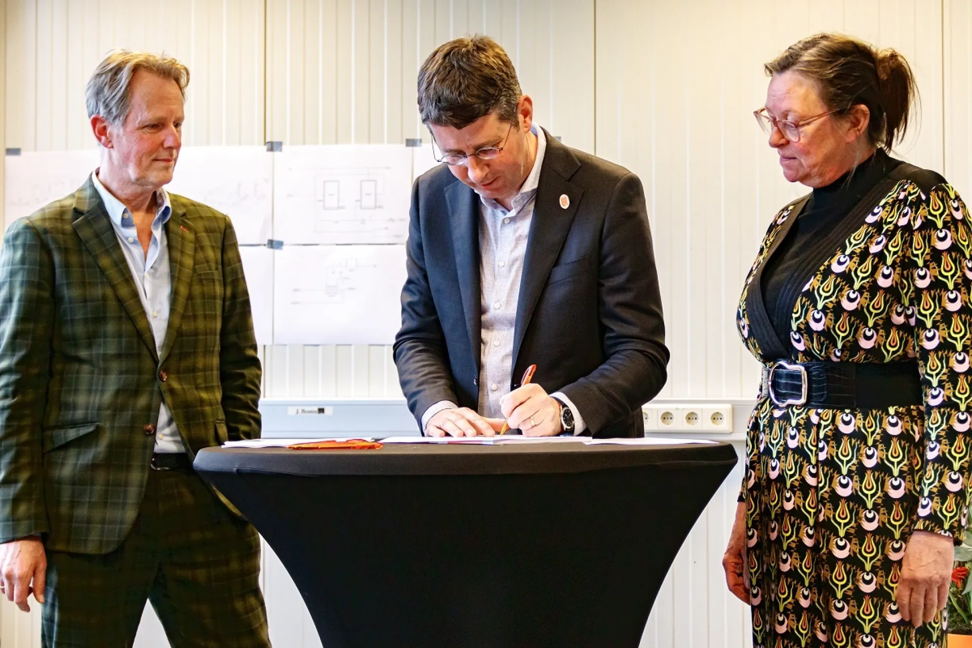 Drie personen bij een hoge tafel terwijl een officieel document wordt ondertekend.