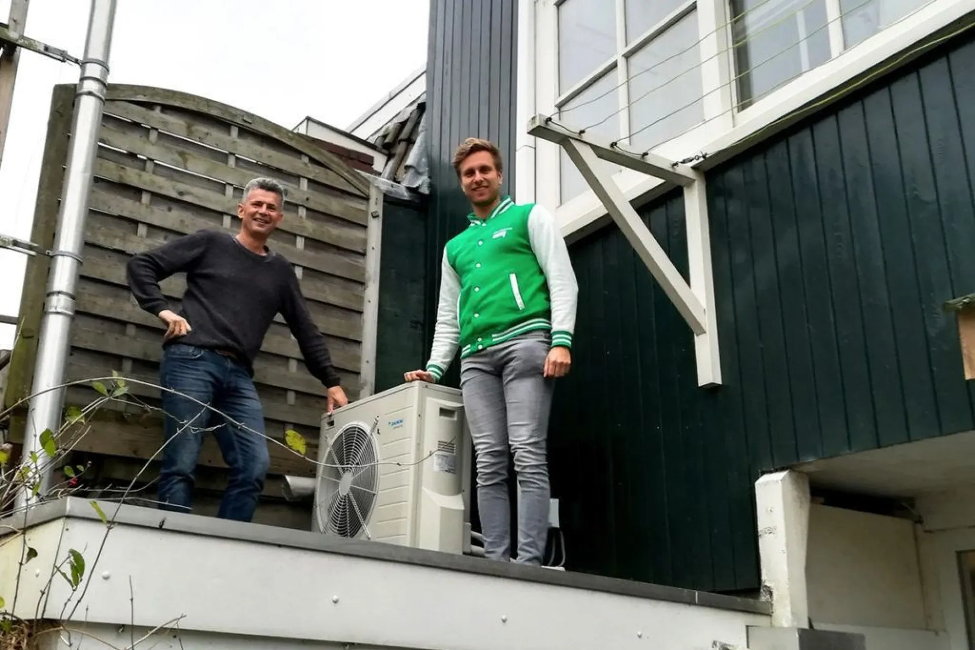 Twee mannen poseren naast een warmtepompunit aan de buitenkant van een huis.